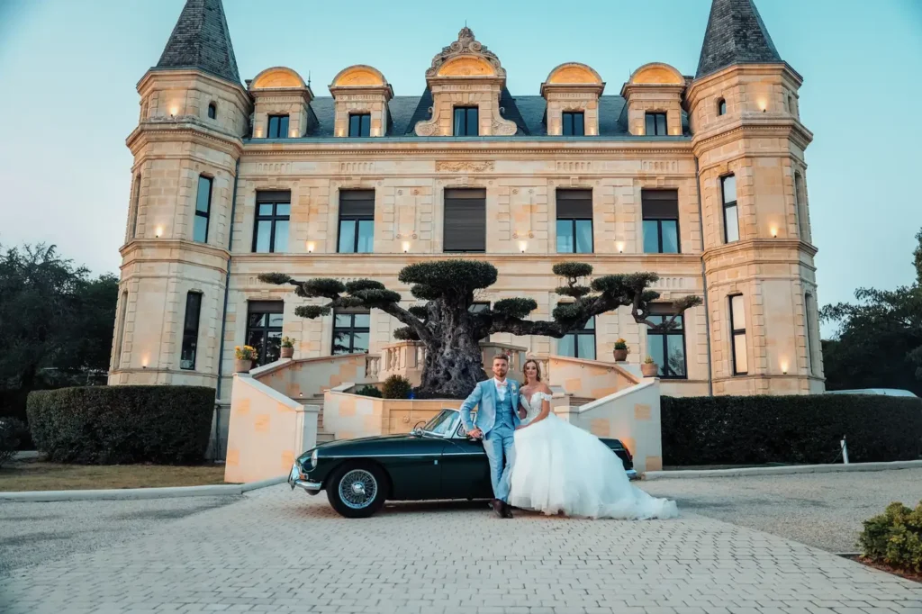 Mariés posant devant le château avec voiture ancienne