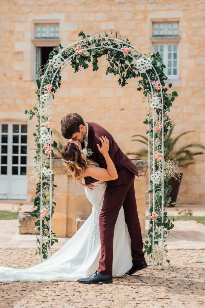 Échange de baiser sous l’arche florale du mariage en Gironde